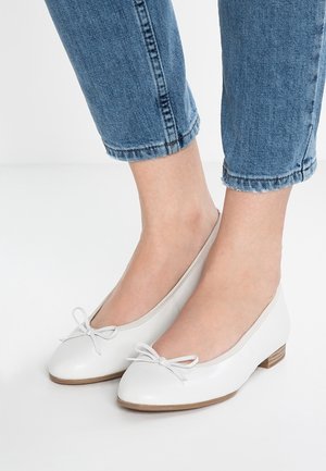 Fødder iført hvide balletsko med lav hæl og sløjfer, kombineret med ankel-lange blå denimjeans mod en lys ens baggrund.
