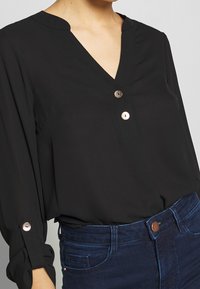 Chemisier noir à manches trois-quarts avec décolleté en V et détails plissés. Présente des boutons ronds en tons dorés et argentés. Associé à un jean bleu.