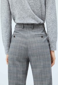 Szare w kratę spodnie z wysokim stanem, z tylnymi kieszeniami z klapami i brązowymi guzikami. Częściowo widoczny szary sweter dzianinowy.