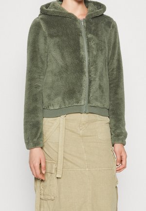 Frau trägt eine olivgrüne, flauschige Kapuzenjacke mit Reißverschluss, Kapuze und langen Ärmeln, kombiniert mit einem beigen Cargo-Rock mit mehreren Taschen.