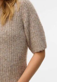 Gros plan d'une personne portant un pull en maille court à manches courtes, de couleur marron clair, avec une texture côtelée.