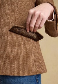 Main tirant un mouchoir de poche marron foncé à motifs de la poche poitrine d'un blazer marron en chevrons sur un jean bleu.