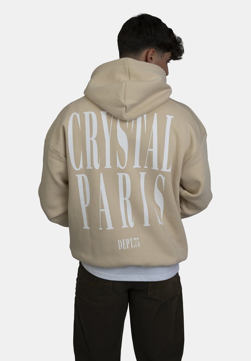 Beige hoodie met witte tekst "CRYSTAL PARIS" en "DEPT.75" op de achterkant. Het materiaal lijkt zacht en gestructureerd, met een oversized ontwerp.