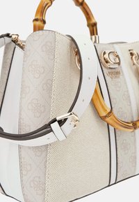 Beżowa torba z białymi akcentami, teksturowana powierzchnia, złote elementy i uchwyt z bambusa. Posiada wzorzysty design oraz regulowany pasek na ramię.