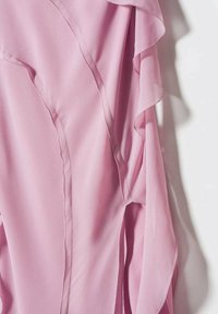 Blouse rose clair en tissu doux et fluide avec des détails volantés le long du bord des manches, présentée sur fond blanc.