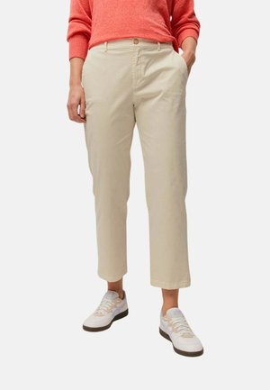 Persona que lleva pantalones beige cortos, zapatillas blancas y un jersey coral de manga larga, de pie con una mano en el bolsillo.