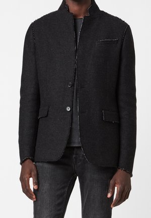 Homme portant un blazer texturé charbon foncé avec deux boutons, un revers sans encoche, des poches plaquées et des coutures apparentes, associé à un jean foncé.