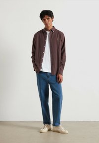 Camicia button-up marrone scuro con una maglietta bianca sotto, abbinata a jeans azzurri chiari e sneakers color crema. Tessuto strutturato, vestibilità casual.