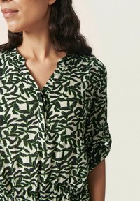 Blusa estampada con un patrón de hojas verdes y negras sobre un fondo claro, con escote en V y cintura fruncida, mangas cortas.