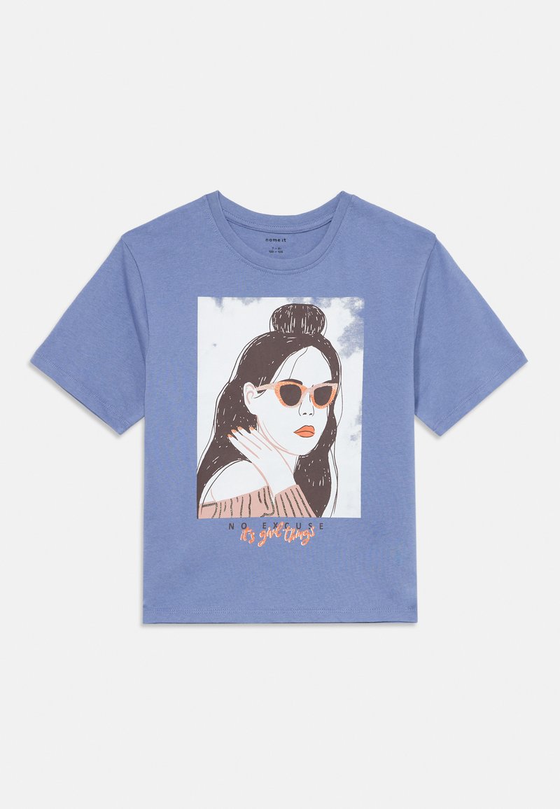 Zila īsām piedurknēm kreklā ar grafiku, kurā attēlota sieviete ar saulesbrillēm un bunā, zem kura ir teksts "Nav attaisnojuma, tā ir meiteņu lieta".