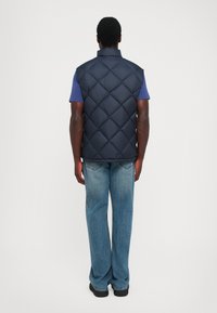 Homme portant un gilet matelassé bleu marine sur une chemise à manches courtes bleue avec un jean bleu clair et des chaussures noires, se tenant de dos.