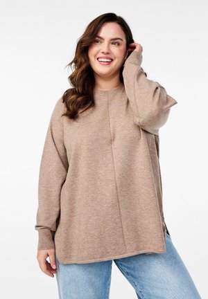 Hellbrauner oversize Pullover mit langen Ärmeln, gerippten Bündchen und seitlichen Schlitzen. Weiche Textur, minimale Nahtdetails, kombiniert mit hellblauen Jeans.