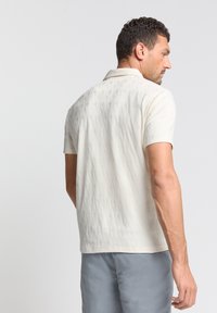 Korte mouwen polo shirt in crème met een gestructureerd patroon, voorzien van een klassieke kraag en een ontspannen pasvorm. De achterkant toont subtiele geometrische ontwerpen.
