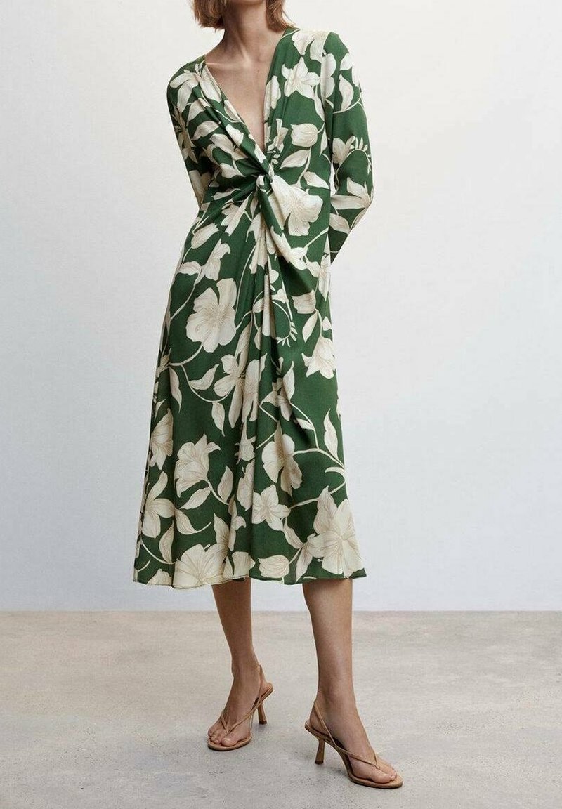Femme portant une robe midi verte à imprimé floral blanc, manches longues, détail noué à l'avant, et sandales à talons beiges, debout sur un sol uni.