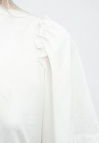 Hvit langermet topp med samlet stoffdetalj på skulderen, laget av myk bomull, med en avslappet passform og glatt tekstur.