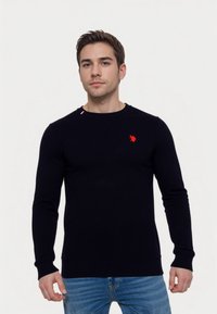 Temno moder pulover z dolgimi rokavi, rebrastimi manšetami in obrobo. Ima majhen rdeč emblem na prsih in kontrastni rdeči poudarek na rami.