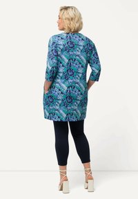 Tunika a fantasia in tonalità di blu e verde con scollatura rotonda, maniche a tre quarti e tasche, abbinata a leggings scuri e tacchi block.