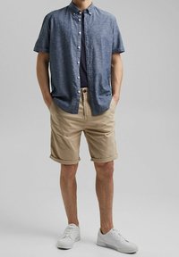 Korte mouwen, donkerblauw button-up shirt over een marineblauw T-shirt, gecombineerd met opgerolde beige shorts en witte sneakers. Glad materiaal, casual ontwerp.