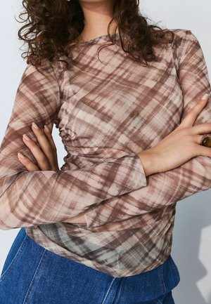 Femme portant un haut à manches longues à carreaux marron et beige avec un jean bleu, bras croisés et une bague à la main droite.