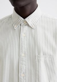 Light green and white striped shirt with a button-down collar, featuring pearl buttons and a chest pocket with embroidered branding.