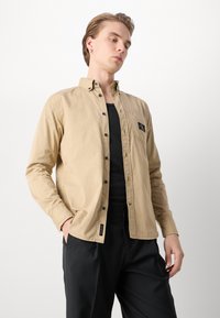 Camicia a maniche lunghe beige chiaro con colletto a bottoni, bottoni neri e tasca sul petto con logo. Indossata su una maglietta nera.