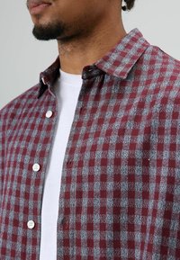 Kariertes Flanellhemd in Burgunderrot und Grau mit Button-Down-Kragen und weißen Knöpfen, getragen über einem weißen T-Shirt.