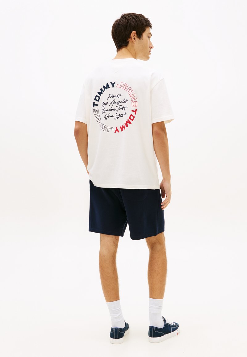 Άνδρας που φοράει λευκό T-shirt Tommy Jeans με κυκλικά ονόματα πόλεων στην πλάτη, σκούρες μπλε βερμούδες, λευκές κάλτσες και σκούρα μπλε αθλητικά παπούτσια, στέκεται πλευρικά.