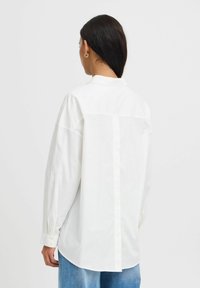Camicia bianca con bottoni, caratterizzata da maniche lunghe, un colletto classico e una cucitura posteriore dritta. Realizzata in un tessuto morbido e leggero.