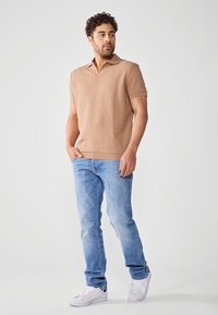 Beige, rövid ujjas póló, texturált mintával, világoskék farmernadrággal és fehér sportcipővel párosítva, egy álló egyénen.