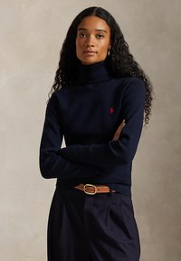 Femme aux longs cheveux bouclés portant un pull à col roulé bleu marine avec un logo rouge, un pantalon foncé et une ceinture en cuir marron, debout les bras croisés.