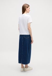 T-shirt blanc à manches courtes associé à une longue jupe en denim. La jupe possède deux poches arrière et une coupe droite. Les chaussures sont des baskets de couleur claire.