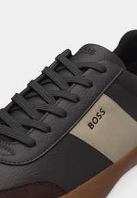 Μαύρο δερμάτινο αθλητικό παπούτσι με σκούρα κορδόνια, μπεζ πλαϊνή επένδυση, καφέ σουέτ λεπτομέρεια και λογότυπο "BOSS" στο πλάι.