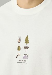 T-shirt blanc en coton avec des motifs de camping illustrés : une lanterne, un arbre, un panneau forestier, une bouilloire et une hache, avec le texte "WILD LIFE."