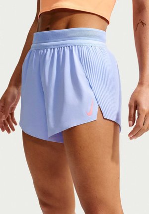 Donna con pantaloncini sportivi viola chiaro con logo Nike rosa e dettaglio laterale plissettato, abbinati a un top corto color pesca.