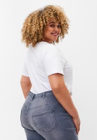 Bela kratka majica s kratkimi rokavi, v kombinaciji z sivo denim hlačami srednje višine, ki imajo zadnje žepke in prilegajočo obliko, na osebi s kodrastimi lasmi.