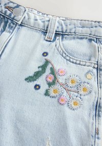 Jean denim bleu clair avec un motif floral brodé en blanc, rose, bleu et vert près de la poche avant et de la boucle de ceinture.