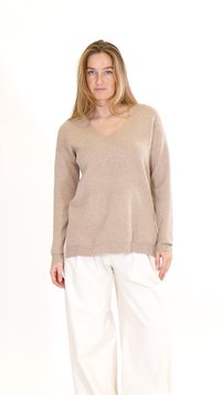 Jauna sieviete ar gariem blondiem matiem, valkājot bēša krāsas V veida kakla džemperi un vaļīgas baltas bikses, stāvot pret baltu fonu.