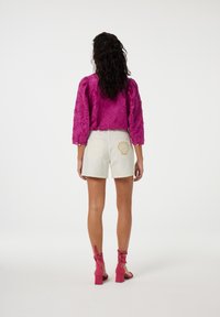 Heldere roze geborduurde blouse met lange mouwen, gecombineerd met witte shorts met schelpborduursel op de achterzak. Roze enkelsandaaltjes met bandjes.