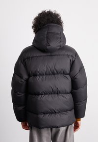 Schwarze Daunenjacke mit Kapuze, die über gesteppten Bereichen und einer glatten Außenseite verfügt. Der Träger ist abgewandt, sodass die Rückendetails und die Ärmel zu sehen sind.