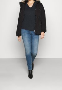 Black parka with a fur-lined hood, paired with a dark blue knit top and light blue jeans, featuring a slim fit and ankle-length black boots.