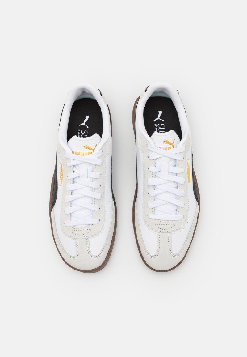 Zapatilla blanca con acentos de ante gris, puntera redonda, cordones planos, suela de goma marrón y interior negro con detalles del logo y marca amarilla.