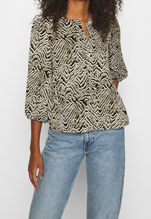 Woman wearing a green and white zebra-striped button-up blouse with 3/4 puff sleeves and light blue denim jeans, standing with one hand in pocket.