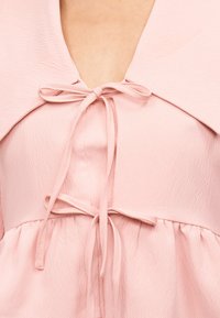 Κοντινό πλάνο ενός ανοιχτό ροζ υφασμάτινου φορέματος με δύο δεμένους φιόγκους στο στήθος και τη μέση, με βαθύ V λαιμόκοψη και ραμμένο ύφασμα κάτω από τη μέση.