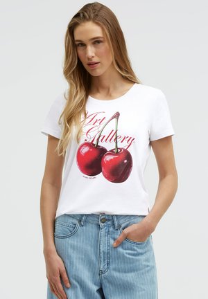 Young woman with long blonde hair wearing a white T-shirt with two large cherries and red "Art Gallery" text, paired with light blue jeans.