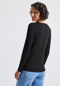 Donna con capelli corti e ricci, vestita con una maglietta nera a maniche lunghe e jeans azzurri chiari, che guarda oltre la spalla contro uno sfondo neutro.