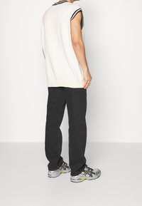 Osoba w bezrękawniku w białym kolorze z czarnym wykończeniem, czarnych dżinsach o luźnym kroju i szarych butach do biegania z żółtymi akcentami, stojąca bokiem.