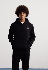 Felpa nera con tasca frontale, cappuccio con coulisse e dettaglio del logo sul petto. Modello che indossa jeans scuri su uno sfondo bianco neutro.