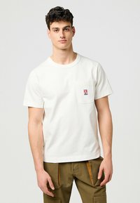 Cremefarvet t-shirt med korte ærmer og rund halsudskæring, der har en brystlomme og et lille mærke. Lavet af blødt bomuldsstof.