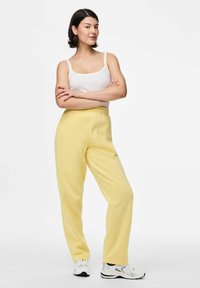Vrouw staat met crossed arms, gekleed in een witte tanktop, gele sweatpants en witte sneakers tegen een effen witte achtergrond.