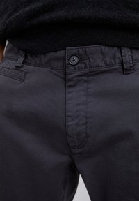 Primer plano de pantalones gris oscuro con cinturilla abotonada y trabillas, combinados con una blusa de punto negra.